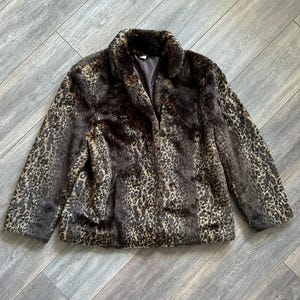 May include: A long-sleeved jacket made of faux fur featuring a brown and black leopard print pattern. The garment has a classic collar, a button-front closure, and a dark brown interior lining visible at the neckline. The texture is thick and plush.
