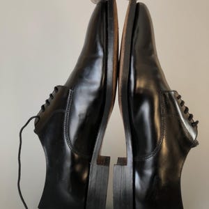 May include: A pair of black leather dress shoes, shown side-by-side. The shoes have a polished, glossy finish and a traditional design. The soles are a contrasting brown colour. The shoes are suitable for formal wear.