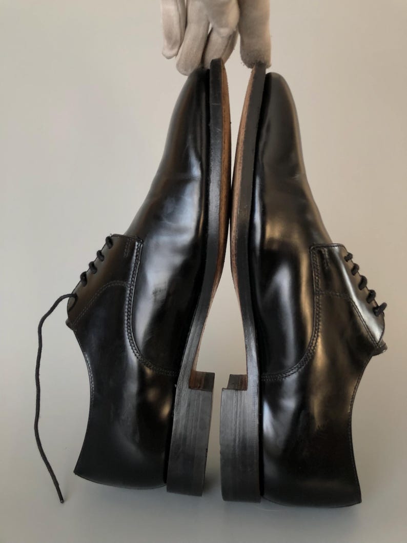 May include: A pair of black leather dress shoes, shown side-by-side. The shoes have a polished, glossy finish and a traditional design. The soles are a contrasting brown colour. The shoes are suitable for formal wear.
