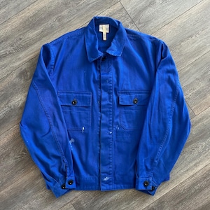 May include: A bright blue long-sleeve work jacket featuring a classic collar, a button-up front, and two chest pockets with button closures. The jacket has long sleeves with buttoned cuffs and displays small white paint splatters on the front.