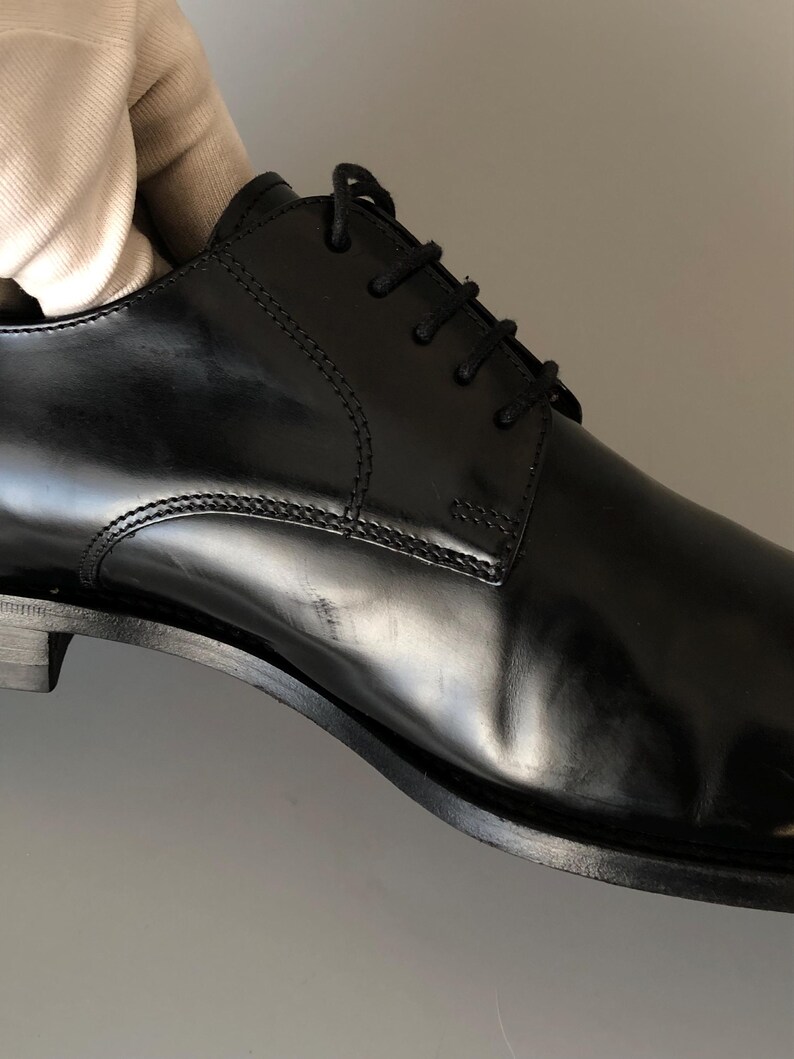 May include: A black leather dress shoe with a polished finish. The shoe has a lace-up design and a low heel. Stitching is visible along the seams. The shoe is a classic style, suitable for formal events. The shoe's length is approximately 28 cm.