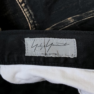 Puede incluir: Primer plano de unos pantalones vaqueros negros con un aspecto descolorido y desgastado. Los pantalones tienen un forro interior blanco y una etiqueta con el texto "Yohji Yamamoto POUR HOMME" en gris.