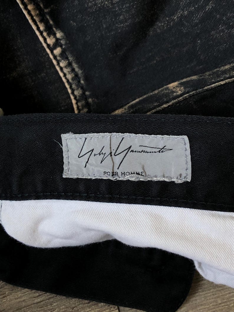 Puede incluir: Primer plano de unos pantalones vaqueros negros con un aspecto descolorido y desgastado. Los pantalones tienen un forro interior blanco y una etiqueta con el texto "Yohji Yamamoto POUR HOMME" en gris.