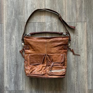 May include: A brown leather shoulder bag featuring a slouchy silhouette, a long adjustable strap, and a shorter top handle. The front displays two flap pockets, one additional zippered pocket, and side lace-up detailing with silver-toned hardware.