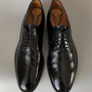 May include: A pair of black leather dress shoes with laces. The shoes have a polished finish and are displayed on a light grey surface. Shoe trees are inserted inside the shoes.