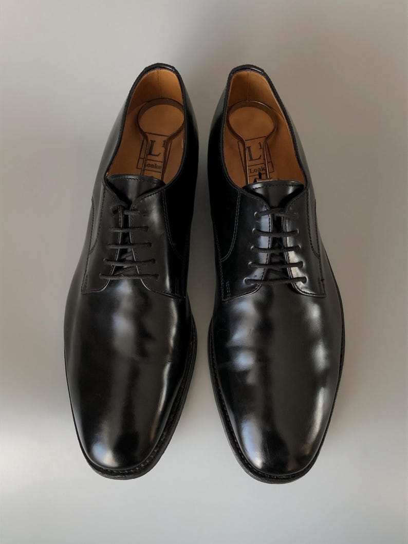 May include: A pair of black leather dress shoes with laces. The shoes have a polished finish and are displayed on a light grey surface. Shoe trees are inserted inside the shoes.