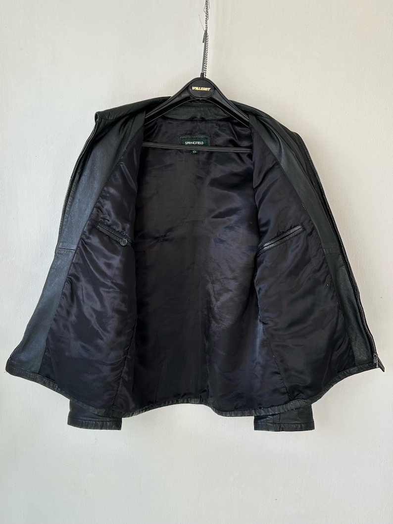 May include: A black leather jacket with a smooth black interior lining, a front zipper closure, and two interior chest pockets. The jacket features a label at the neck with the text Springfield, and it is displayed on a black plastic hanger.