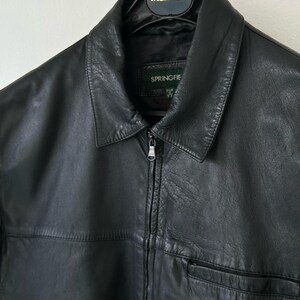 May include: A black leather jacket featuring a classic collar, a full-length front zipper closure with a metal pull, and a horizontal seam across the chest. The interior label displays the brand name Springfield, and it hangs on a black hanger.