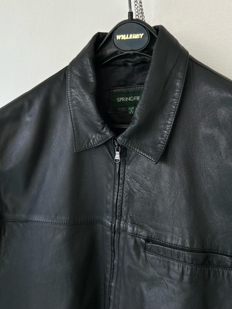 May include: A black leather jacket featuring a classic collar, a full-length front zipper closure with a metal pull, and a horizontal seam across the chest. The interior label displays the brand name Springfield, and it hangs on a black hanger.