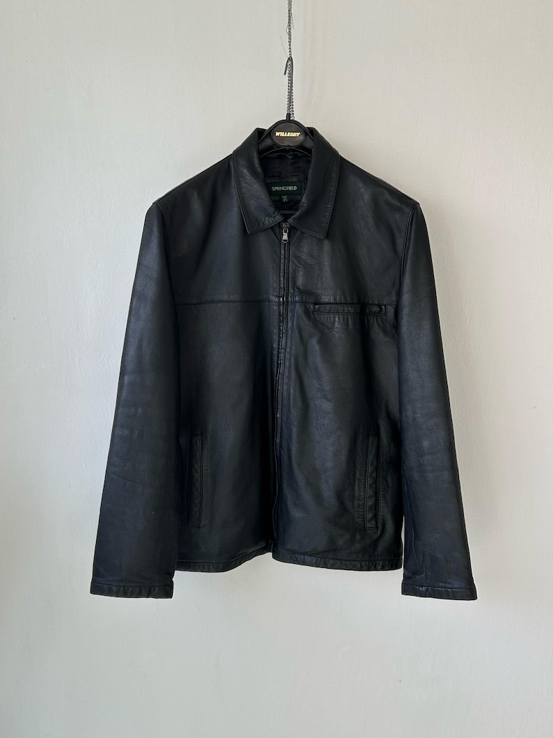 May include: A black leather jacket featuring a classic collar, a full-length front zipper closure, and two lower side pockets. The garment includes a horizontal seam across the chest and a small chest pocket, with a Springfield brand label inside.