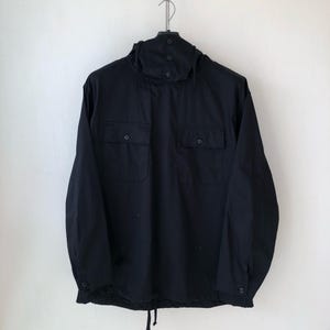May include: A navy blue anorak-style jacket with a hood and two front pockets. The jacket has a drawstring at the bottom hem and buttons on the hood and cuffs. The jacket is hanging from a metal hanger.