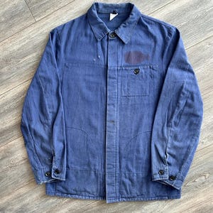 May include: A blue work jacket featuring a collared neckline, a button-down front, and long sleeves with buttoned cuffs. The garment includes a single chest pocket with a button closure and a dark circular patch detail on the upper right chest area.