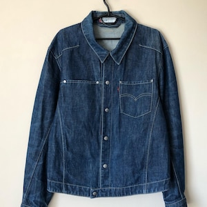 Puede incluir: Chaqueta vaquera azul oscuro con cierre de botones, dos bolsillos en el pecho y una pequeña etiqueta roja en el bolsillo derecho. La chaqueta está colgada de una percha negra. Cuello clásico y mangas largas.