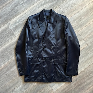 May include: A double-breasted blazer in a dark navy blue satin finish. The garment features wide lapels, four front buttons arranged in two rows, two flap pockets at the waist, and three small buttons on each sleeve cuff with a structured collar.
