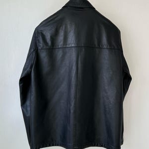 May include: A black jacket with a smooth texture, featuring a classic collar, long sleeves, and a horizontal seam across the upper back. The garment is displayed hanging from a metal hanger, showcasing a clean, structured design with a straight hem.