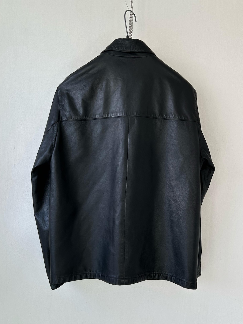 May include: A black jacket with a smooth texture, featuring a classic collar, long sleeves, and a horizontal seam across the upper back. The garment is displayed hanging from a metal hanger, showcasing a clean, structured design with a straight hem.