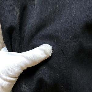 Puede incluir: Primer plano de tela vaquera negra con sutil decoloraci&oacute;n y textura. La tela est&aacute; sostenida por una mano enguantada, mostrando el color oscuro y el tejido del material. El denim parece desgastado.