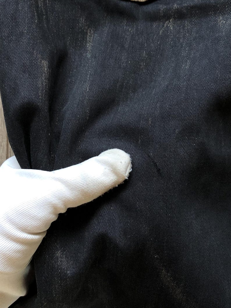 Puede incluir: Primer plano de tela vaquera negra con sutil decoloraci&oacute;n y textura. La tela est&aacute; sostenida por una mano enguantada, mostrando el color oscuro y el tejido del material. El denim parece desgastado.