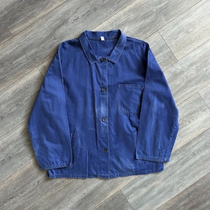 Puede incluir: Una chaqueta de trabajo azul de manga larga con cuello clásico, cierre frontal de botones con cuatro botones visibles y tres bolsillos de parche. La prenda presenta un aspecto desgastado con sutiles variaciones de color en el tejido.