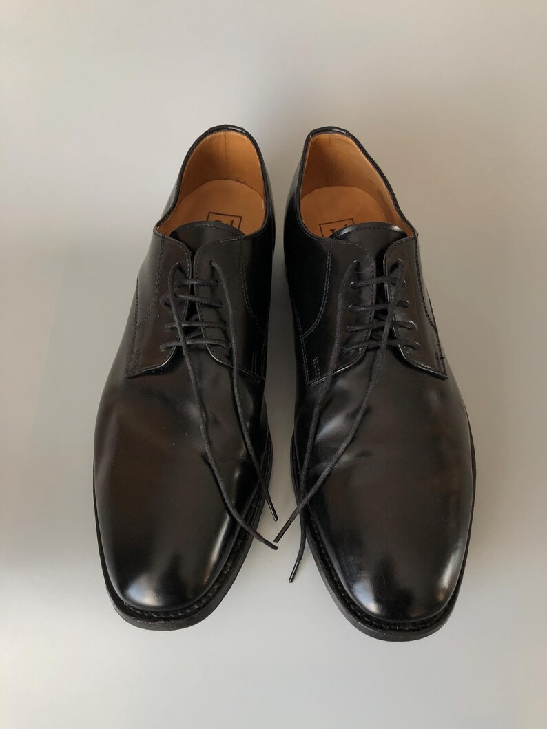 May include: A pair of black leather dress shoes with laces. The shoes have a glossy finish and a classic design, suitable for formal occasions. The soles are dark, and the interior is a light brown colour.