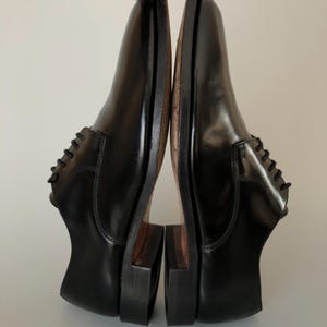 May include: Pair of black leather dress shoes with a polished finish. The shoes feature a traditional lace-up design and a stacked heel. The soles are a contrasting brown colour. The shoes are held up by a gloved hand.