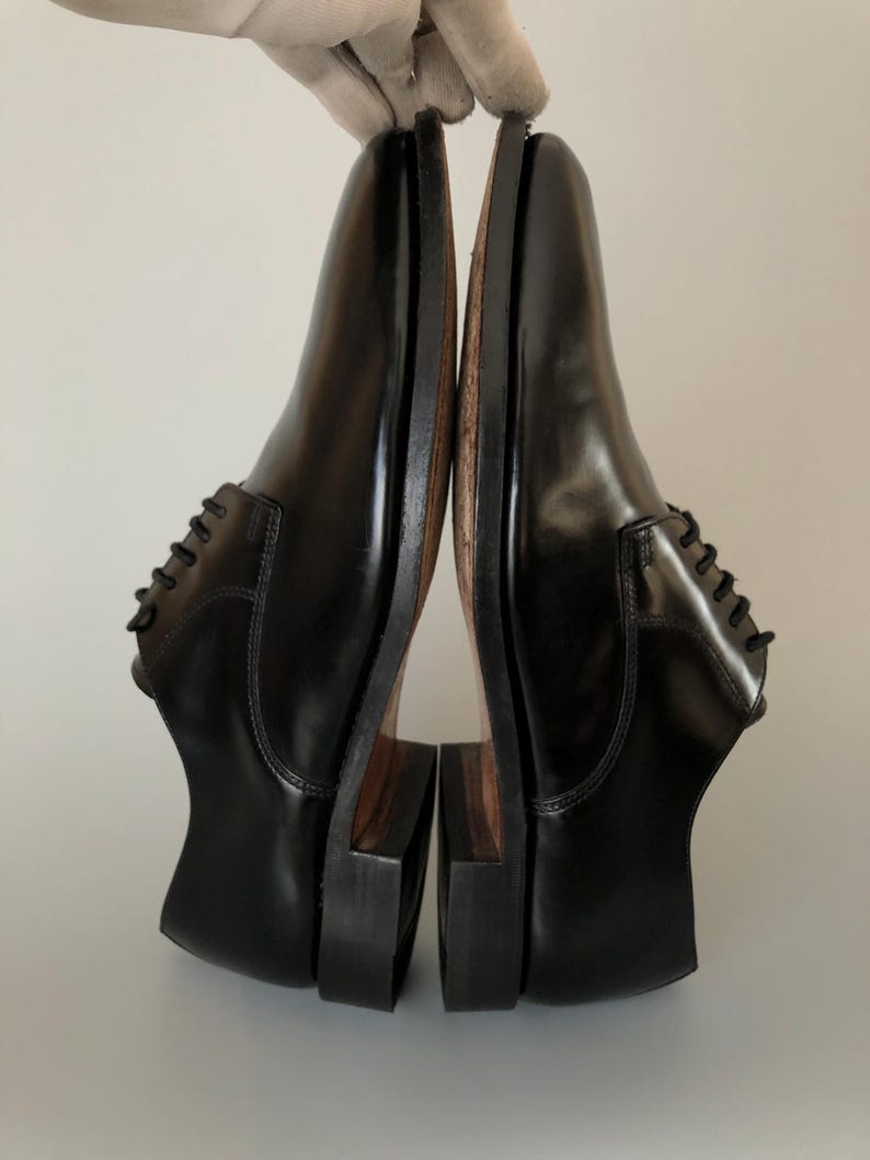 May include: Pair of black leather dress shoes with a polished finish. The shoes feature a traditional lace-up design and a stacked heel. The soles are a contrasting brown colour. The shoes are held up by a gloved hand.