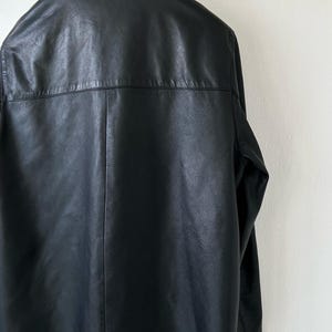 May include: A black leather jacket shown from the back, featuring a classic collar, a horizontal seam across the upper back, and a vertical center seam running down the length of the garment. The jacket has long sleeves and a smooth finish.