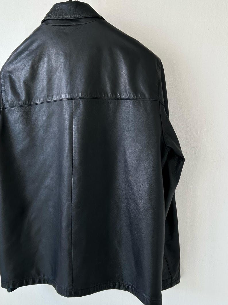 May include: A black leather jacket shown from the back, featuring a classic collar, a horizontal seam across the upper back, and a vertical center seam running down the length of the garment. The jacket has long sleeves and a smooth finish.