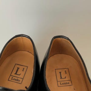 May include: A pair of black leather dress shoes. The insoles are tan and feature the "L1 Loake" logo. The shoes have a classic design with a glossy finish and visible stitching.