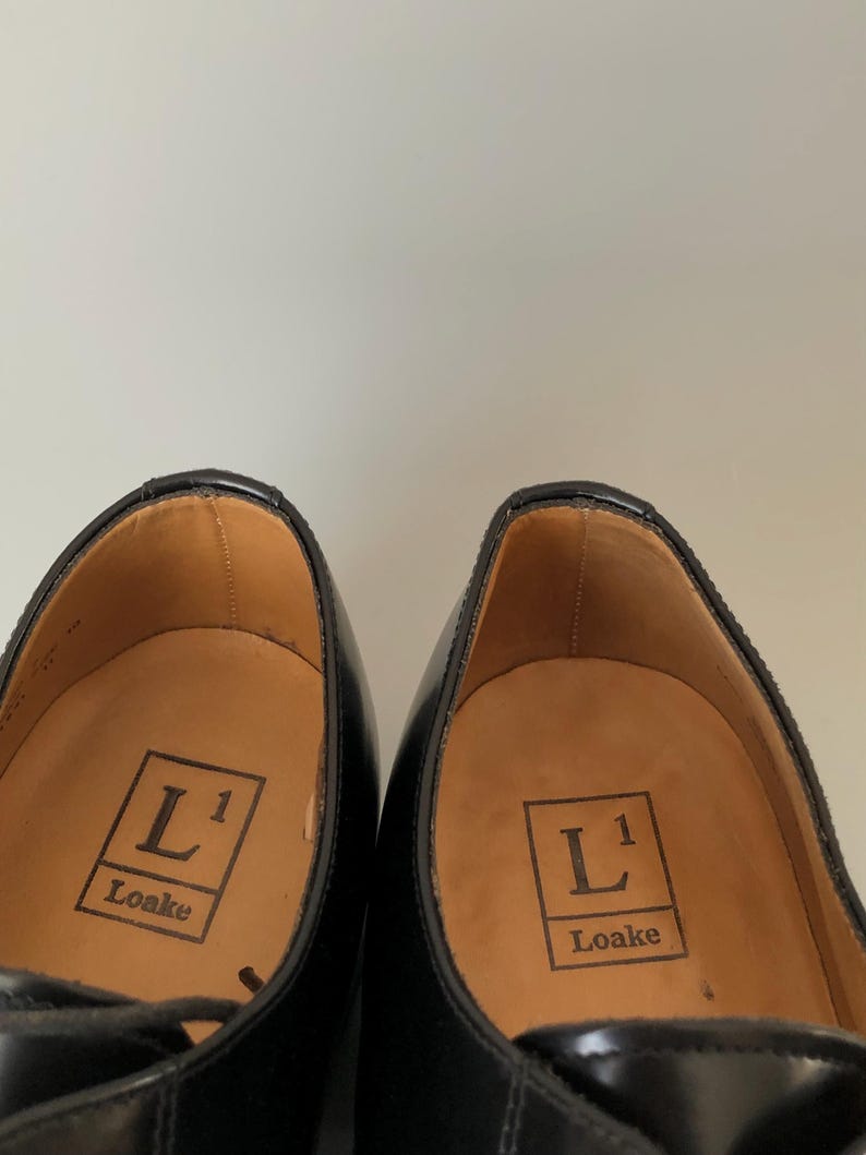 May include: A pair of black leather dress shoes. The insoles are tan and feature the "L1 Loake" logo. The shoes have a classic design with a glossy finish and visible stitching.