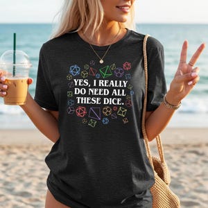Puede incluir: Camiseta gris oscuro con el texto "YES, I REALLY DO NEED ALL THESE DICE." rodeado de formas geom&eacute;tricas coloridas. La camiseta es de cuello redondo.
