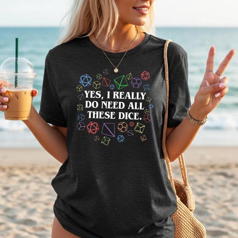 Puede incluir: Camiseta gris oscuro con el texto "YES, I REALLY DO NEED ALL THESE DICE." rodeado de formas geom&eacute;tricas coloridas. La camiseta es de cuello redondo.