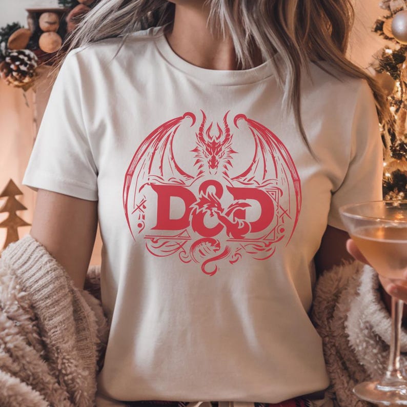Puede incluir: Camiseta blanca con un dise&ntilde;o rojo que presenta un drag&oacute;n y las letras "D&D". El dise&ntilde;o incluye alas de drag&oacute;n y elementos decorativos. La camiseta tiene mangas cortas y cuello redondo.