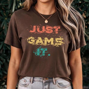 Puede incluir: Una camiseta marr&oacute;n con la frase "JUST GAME IT" en letras coloridas y desgastadas. La camiseta tiene cuello redondo y mangas cortas. El texto es de color naranja, amarillo y turquesa.