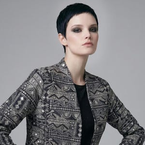 May include: A black and silver patterned jacket with a geometric design. The jacket has long sleeves and is worn over a black top and pleated skirt. The jacket features silver embellishments.