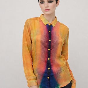 May include: A long-sleeved button-down shirt featuring a vibrant, multi-coloured tie-dye pattern in shades of yellow, orange, red, and blue. The shirt has a pointed collar, a dark blue central placket with yellow buttons, and a fine, vertical pleated texture.