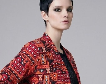 May include: A cropped jacket featuring a vibrant red and orange geometric pattern with integrated small circular mirrors. The jacket has long sleeves, a collarless neckline, and a straight hem, worn over a black top and a black skirt.