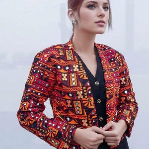 Può includere: Giacca ricamata con scollo a V, con un motivo geometrico rosso, arancione e nero. La giacca ha maniche lunghe ed è decorata con piccoli abbellimenti riflettenti. La modella indossa un top nero sotto.