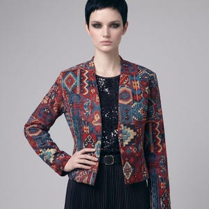Könnte beinhalten: Gemusterte Jacke in Rot-, Blau- und Beigetönen. Die Jacke hat ein geometrisches Design und wird über einem schwarzen Oberteil und einem plissierten Rock getragen. Ein schwarzer Gürtel betont die Taille.