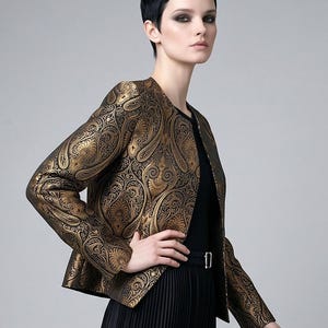 May include: A gold and black paisley patterned jacket with a black top and pleated skirt. The jacket has a classic collarless design and long sleeves. The model is wearing a black belt with a silver buckle.
