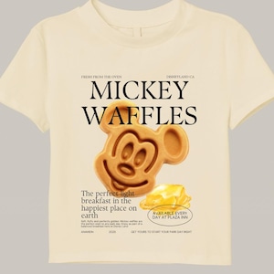 May include: Cream-colored t-shirt featuring a Mickey Mouse waffle design. The graphic includes the words "MICKEY WAFFLES" and text that reads "The perfect light breakfast in the happiest place on earth." The shirt also has the text "AVAILABLE EVERY DAY AT PLAZA INN."