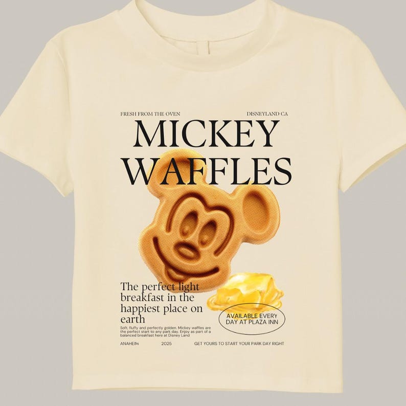 May include: Cream-colored t-shirt featuring a Mickey Mouse waffle design. The graphic includes the words "MICKEY WAFFLES" and text that reads "The perfect light breakfast in the happiest place on earth." The shirt also has the text "AVAILABLE EVERY DAY AT PLAZA INN."