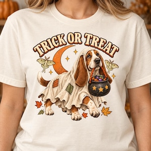 May include: A cream-colored t-shirt featuring a graphic of a basset hound dressed as a ghost with a patchwork sheet. The dog carries a black cauldron filled with candy. Above the dog, the text reads TRICK OR TREAT, with a crescent moon, moths, and autumn leaves surrounding the scene.