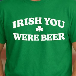 May include: Green t-shirt with the white text "IRISH YOU WERE BEER" and a shamrock graphic. The shirt is a solid green color and is made of a soft material. Perfect for St. Patrick's Day or any casual occasion.