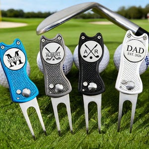 Puede incluir: Cuatro herramientas de reparación de divots de golf personalizadas en azul, gris, negro y plateado, cada una con un diseño y texto únicos. Las herramientas están en un campo de golf verde con pelotas de golf y un palo de golf en el fondo.