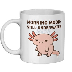 May include: White ceramic mug with a grumpy axolotl illustration and the text "MORNING MOOD: STILL UNDERWATER". The axolotl is pink with orange gills and a displeased expression. Bubbles are drawn above the axolotl.