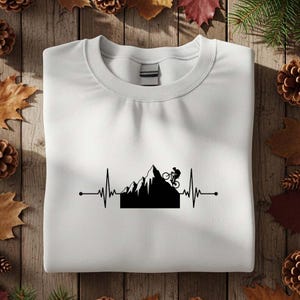 以下が含まれることがあります： 白いスウェットシャツ。黒いグラフィックで山脈、サイクリスト、心拍ラインが描かれています。デザインは中央に配置されています。スウェットシャツは、松ぼっくりと葉のある木製の表面に折りたたまれています。