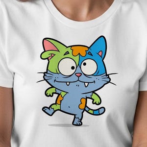 May include: White t-shirt featuring a cartoon cat with a split green and blue head, orange accents, and a playful expression. The cat is light blue with green and orange spots. The design is a whimsical illustration.