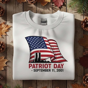 Puede incluir: Sudadera blanca con un gráfico de la bandera estadounidense, las Torres Gemelas y el texto "PATRIOT DAY - SEPTEMBER 11, 2001." El diseño es en rojo, blanco y azul. La sudadera está doblada sobre una superficie de madera.