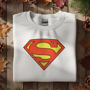 May include: White crew neck sweatshirt featuring the Superman logo. The logo is a red and yellow diamond shape with a distressed, vintage look. The sweatshirt is folded neatly on a wooden surface.